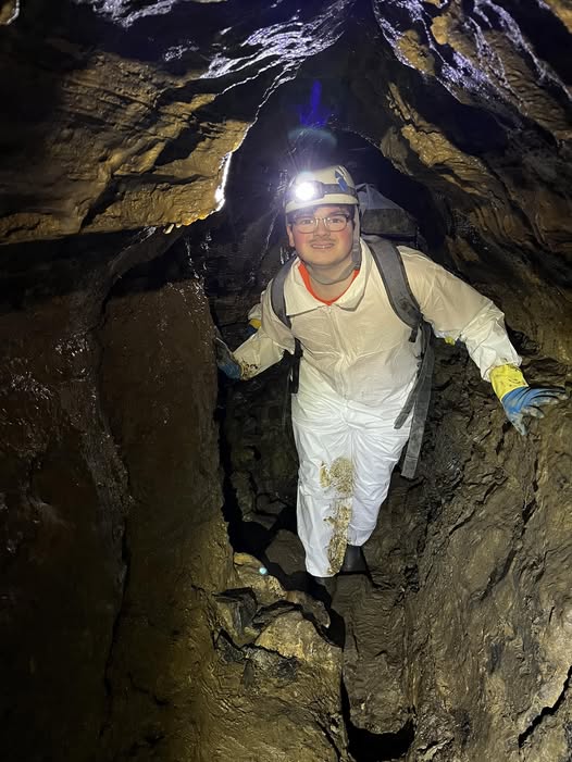 Smiling explorer in a white caving suit, helmet and headlamp squeezing through a narrow, rugged cave passage